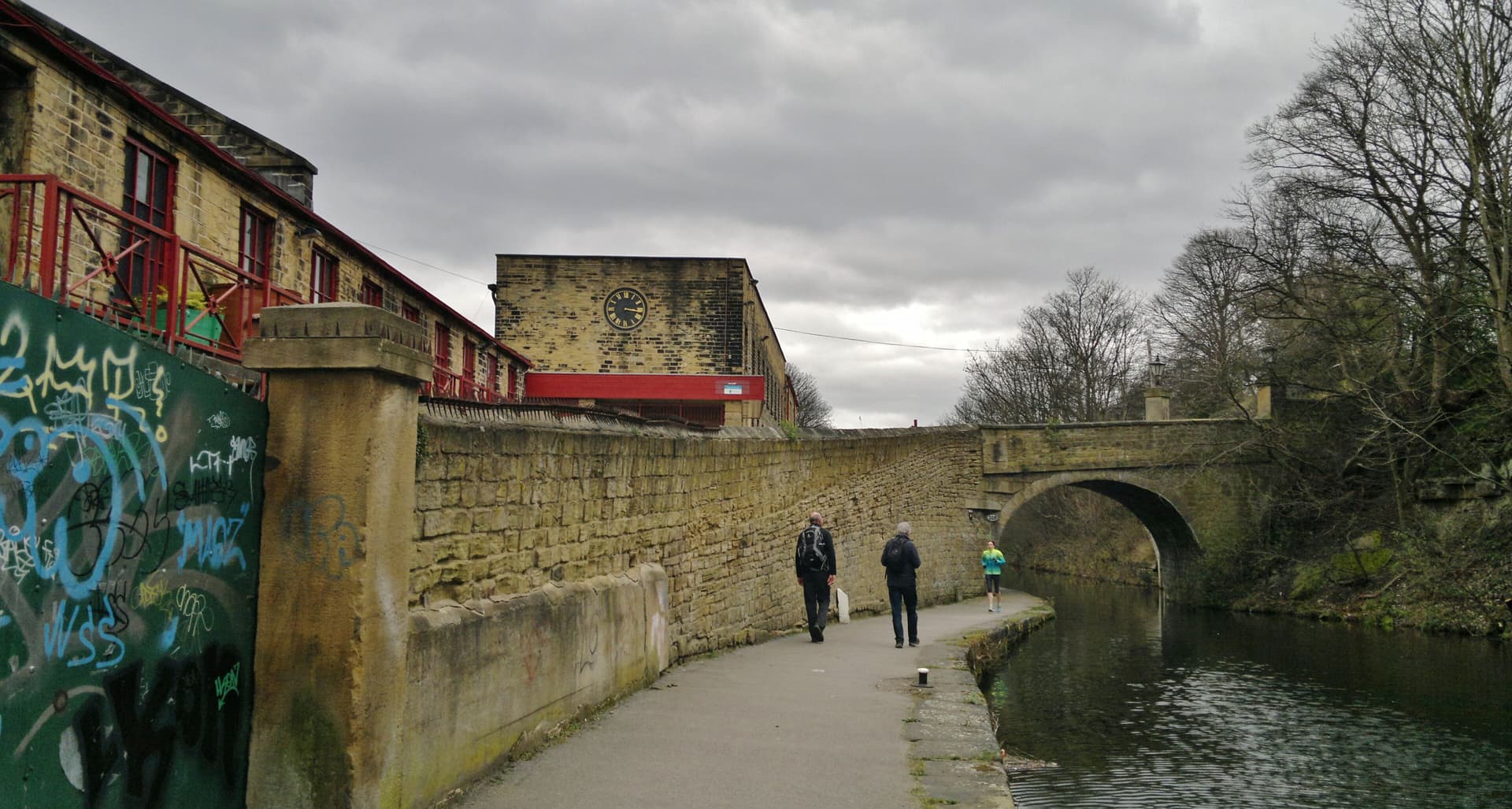 Canal towpath beside a stone wall and old mill buildings with a clock, walkers and a jogger heading towards a bridge