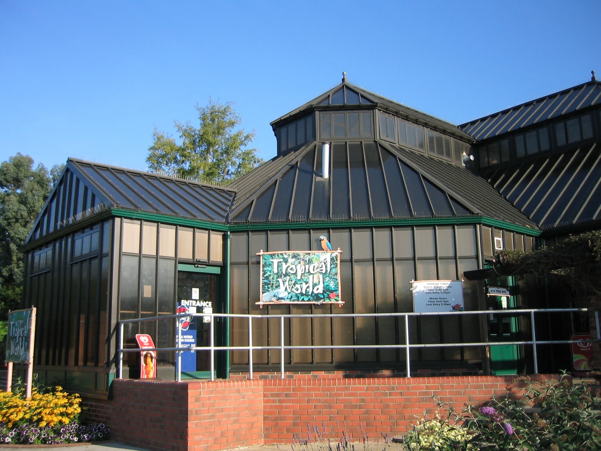 Dark green-framed Tropical World building with a parrot sign, brick wall and flower beds under a clear blue sky