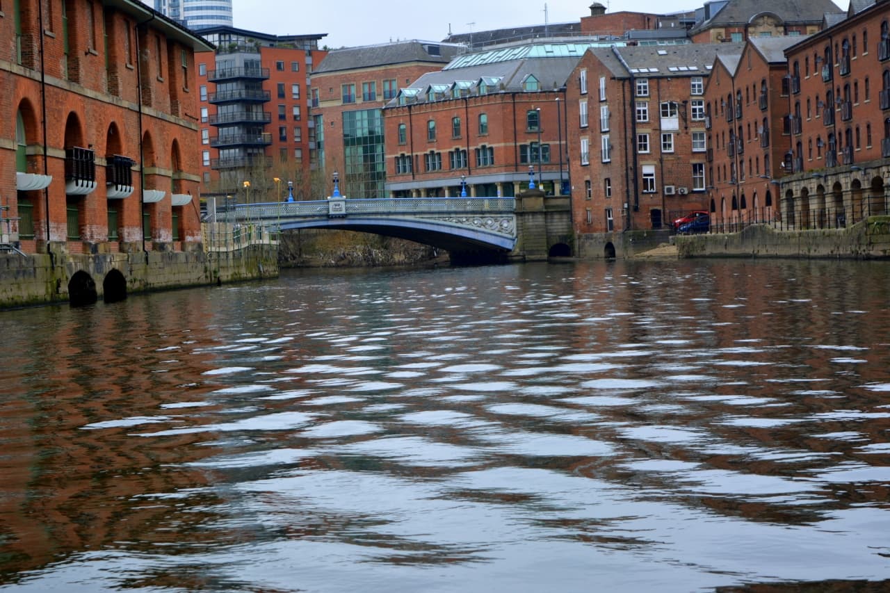 Ornate iron bridge crossing the River Aire, flanked by red-brick warehouses and modern flats under an overcast sky