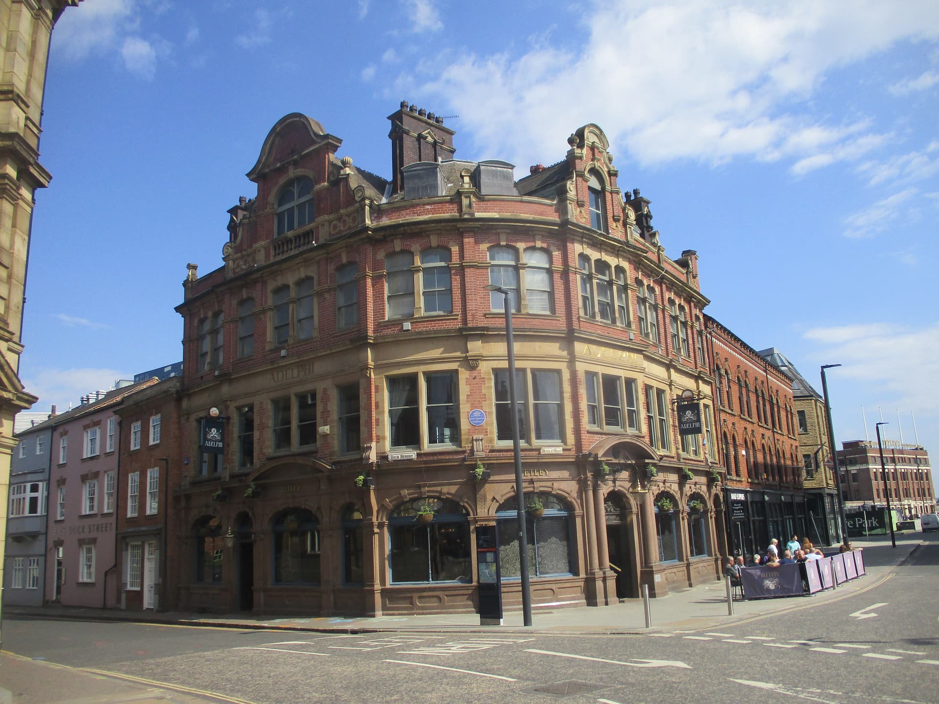 Curved red-brick Victorian corner pub, The Adelphi, with stone detailing and outdoor drinkers under a bright blue sky