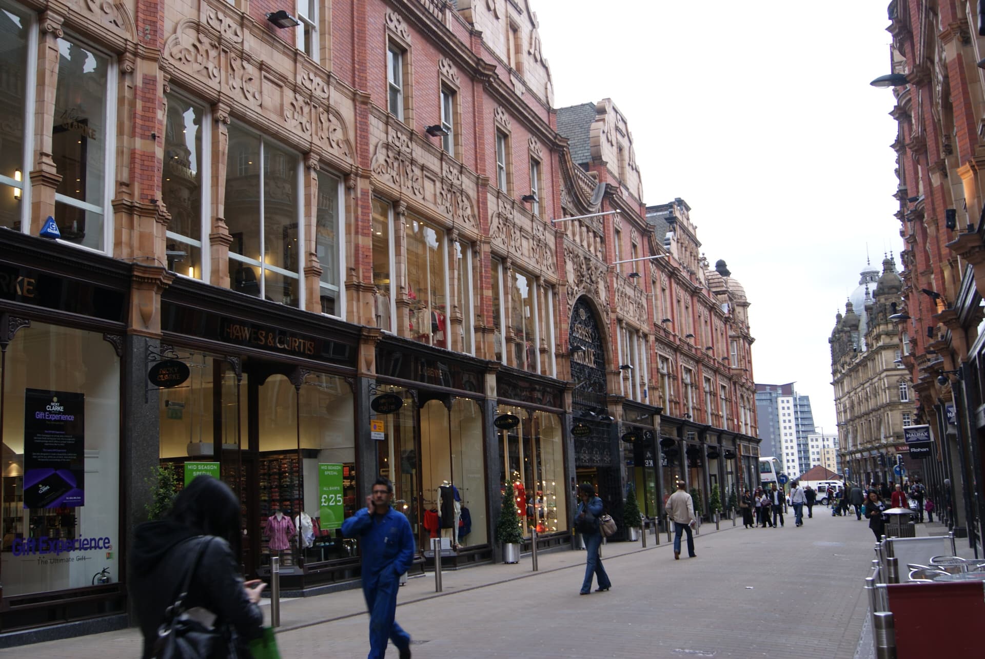 Ornate red-brick and terracotta shopping street with pedestrians, shopfronts and the Cross Arcade entrance in Leeds city centre