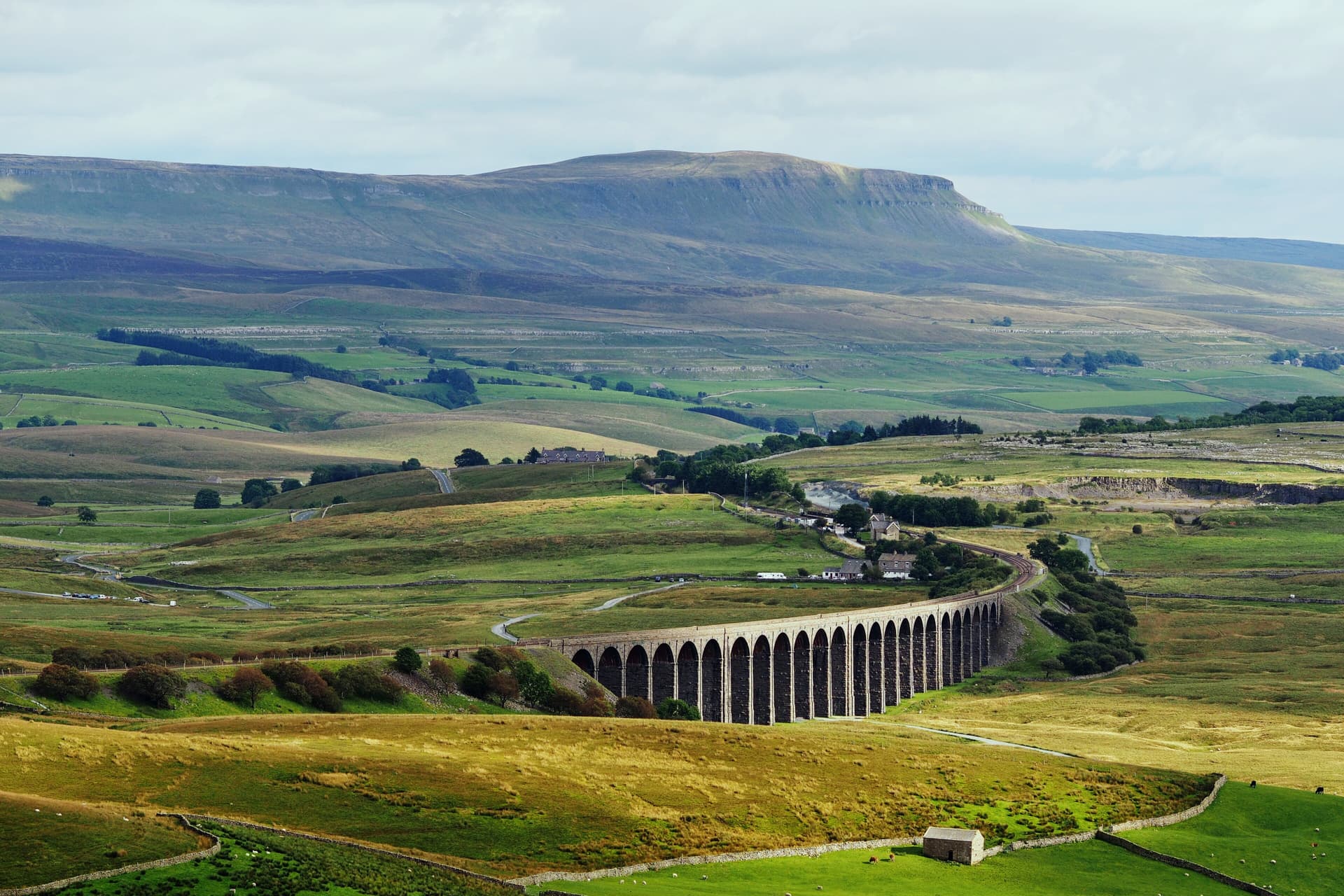 Curving stone railway viaduct crossing green moorland beneath a flat-topped hill in the Yorkshire Dales