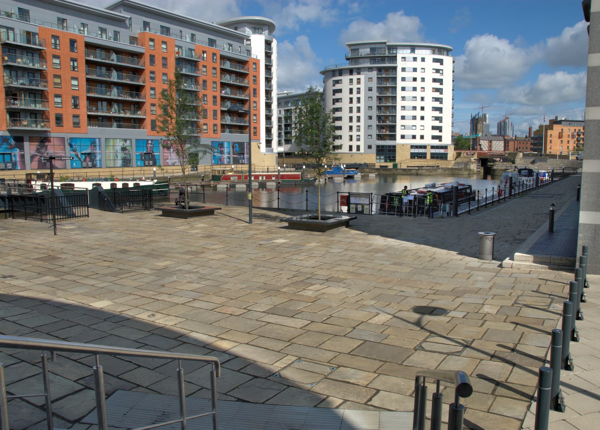 Paved waterfront square beside a Leeds dock basin with moored narrowboats and modern brick and white apartment blocks