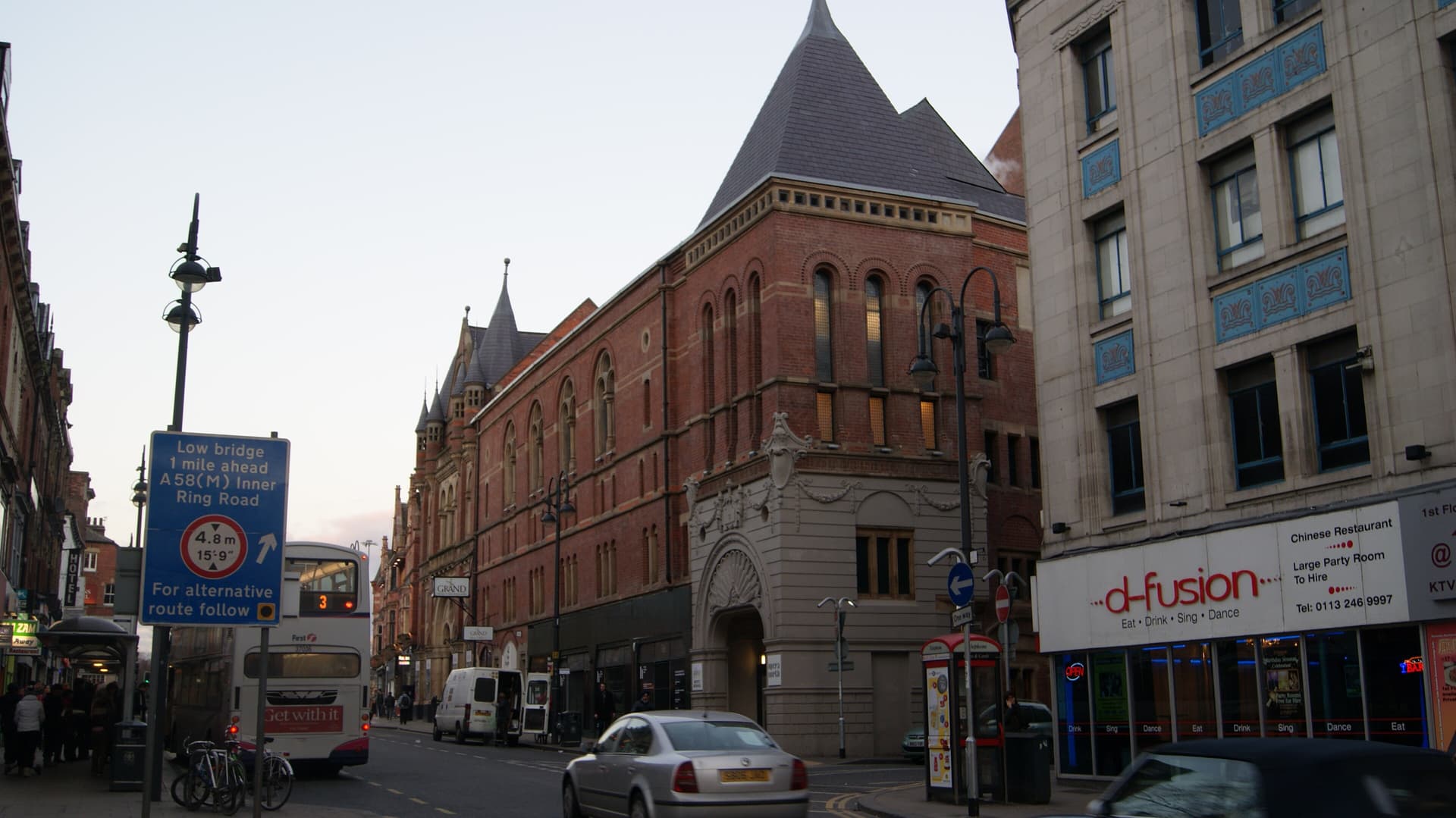 Red-brick Victorian theatre with slate-roofed turrets on a Leeds street corner, buses and cars passing at dusk