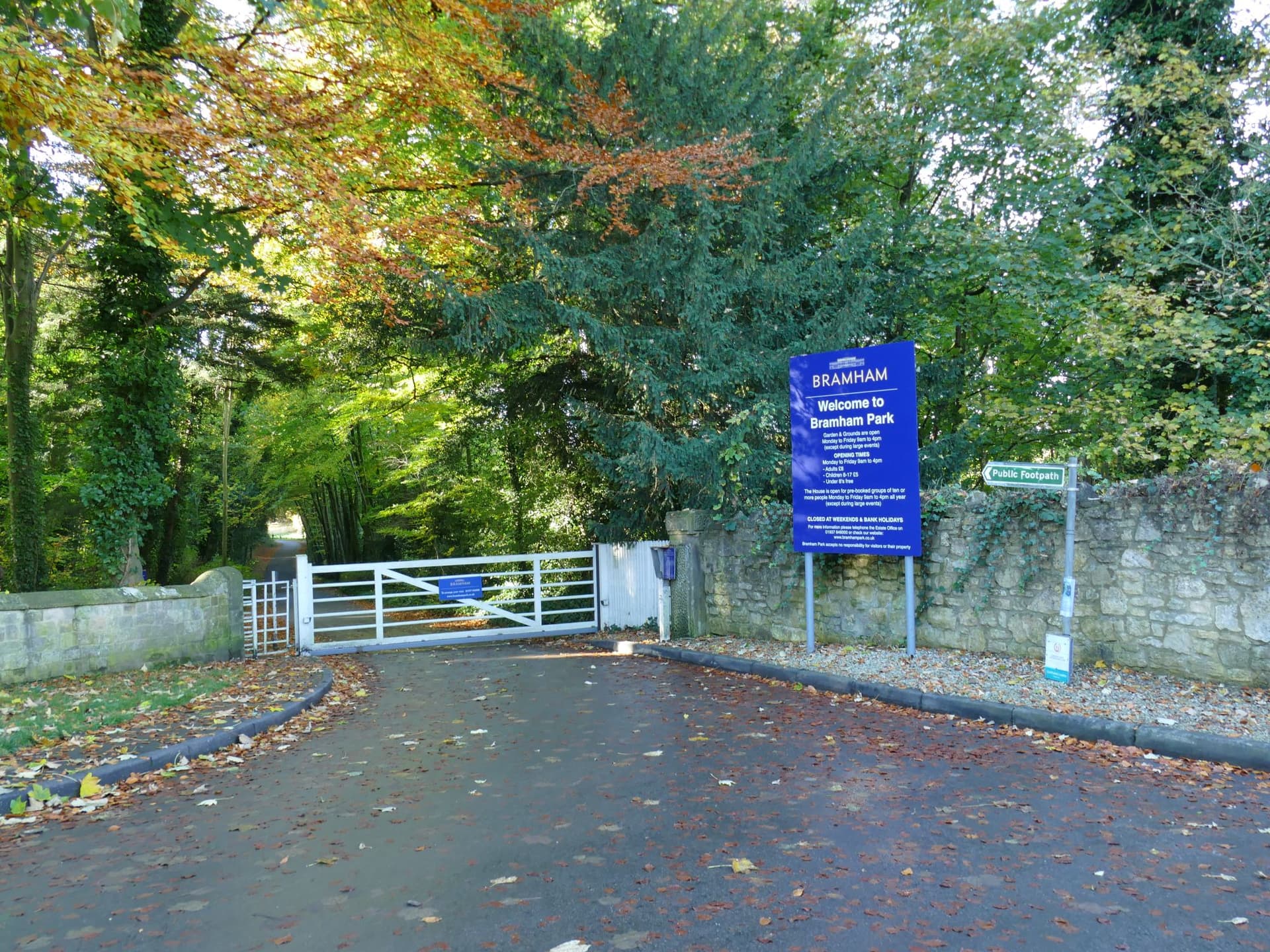 White gate and blue welcome sign at the entrance to Bramham Park, beneath autumn trees