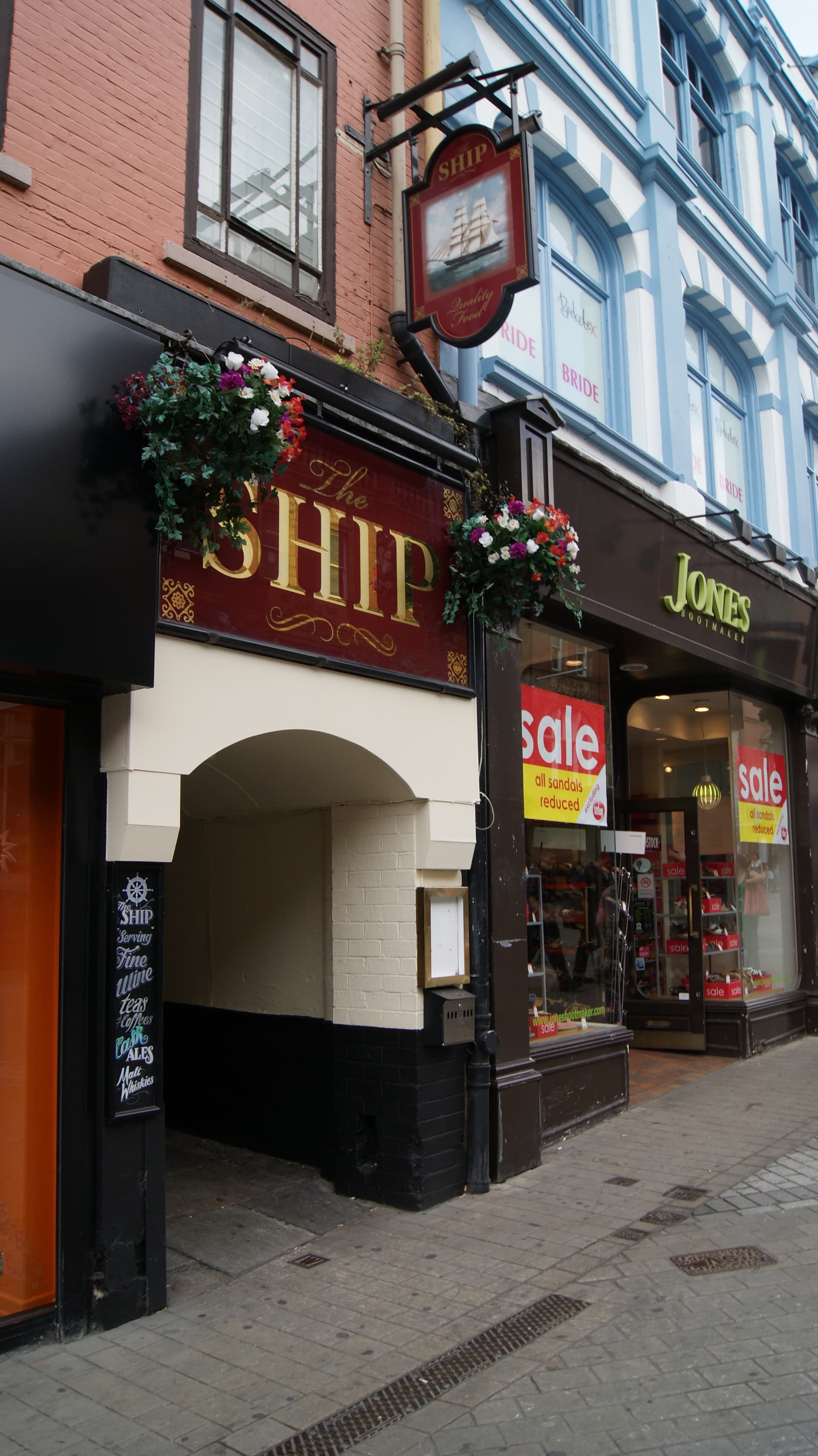 Arched passage entrance to The Ship pub with hanging sign and flower baskets beside a Jones Bootmaker shop