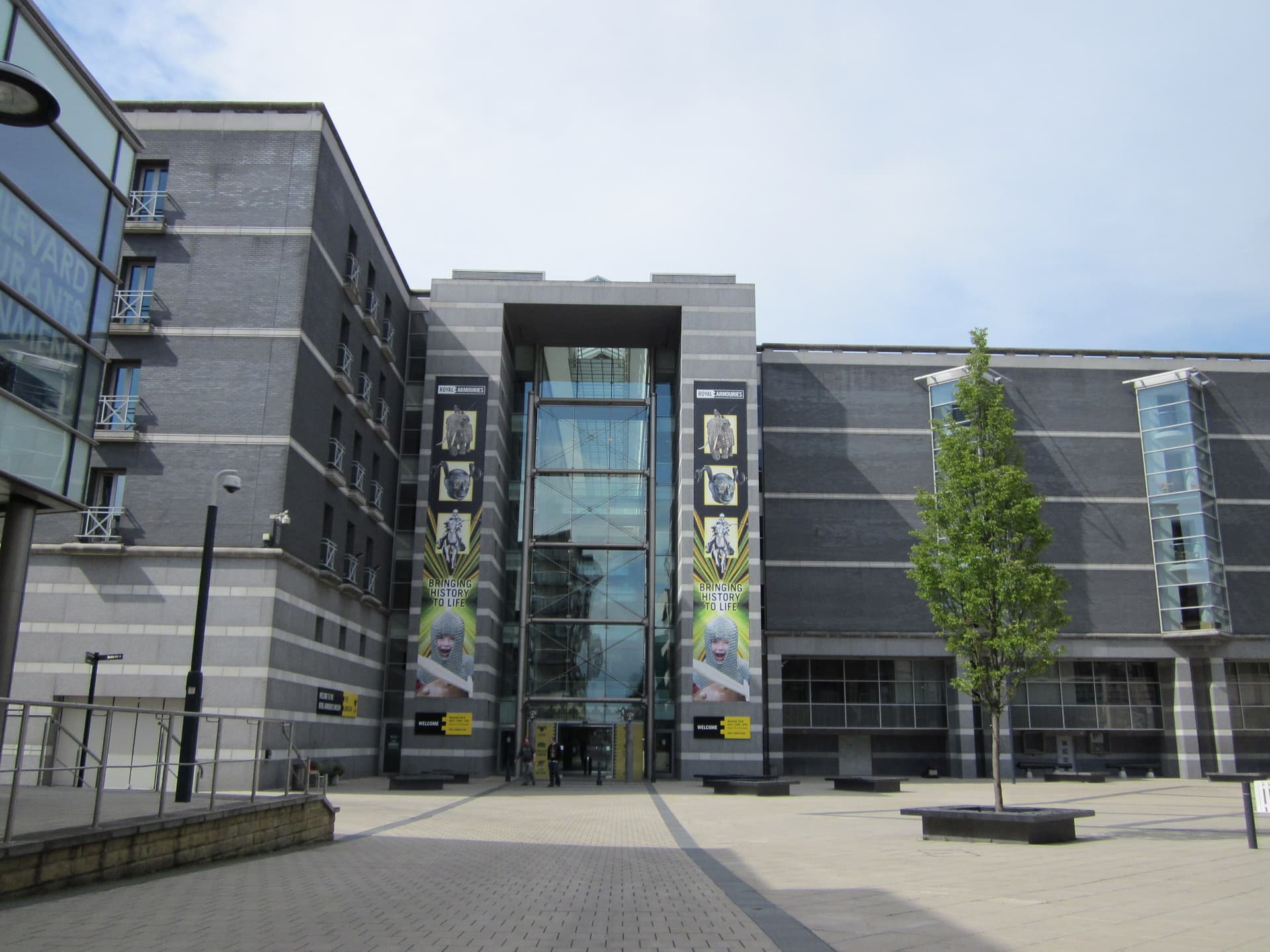 Granite and glass entrance of the Royal Armouries Museum in Leeds, with tall banners and a young tree in the square