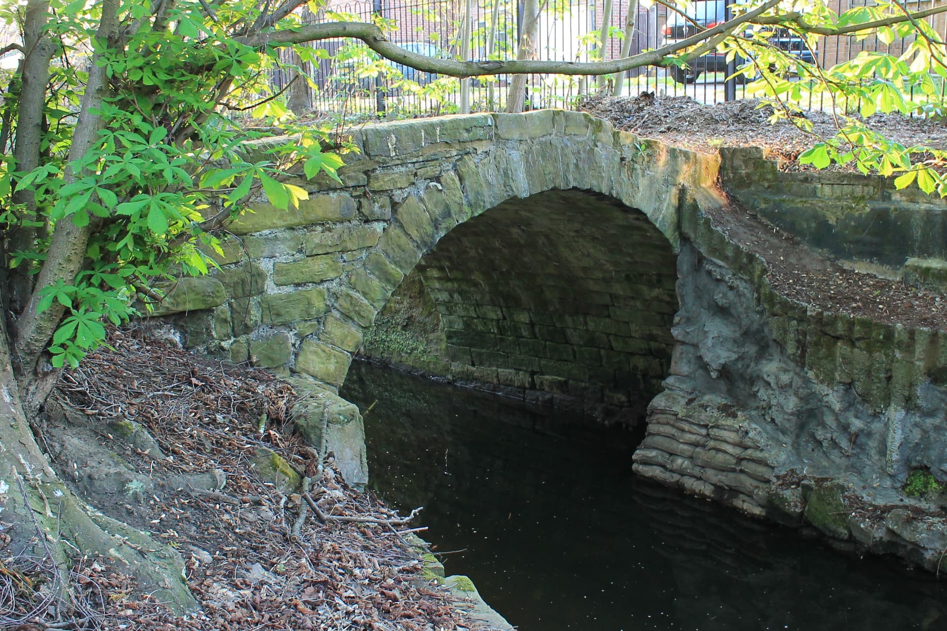 Moss-covered stone arch bridge over dark still water beneath horse chestnut leaves, with railings behind