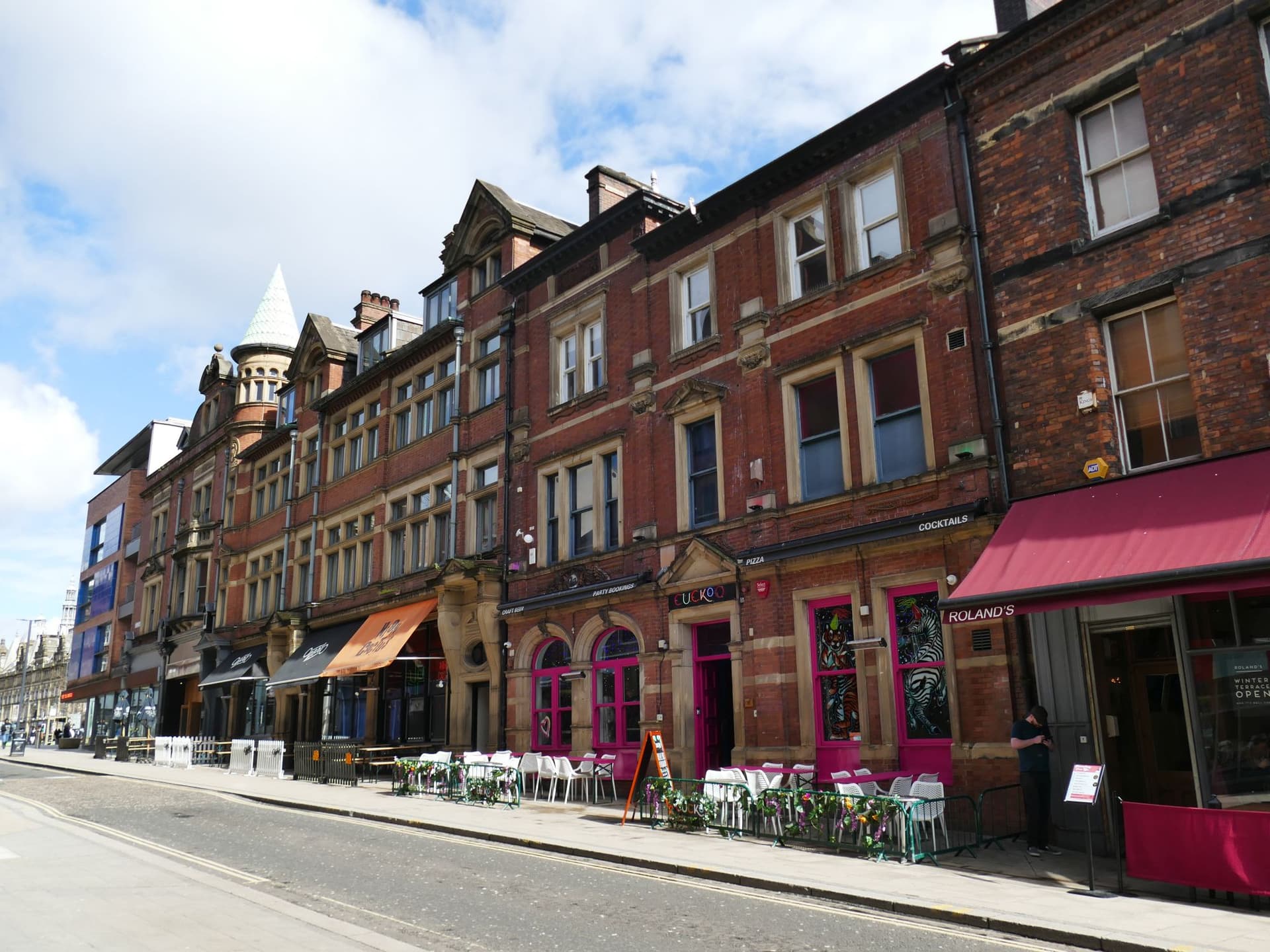 Row of red-brick Victorian buildings with colourful bar frontages and outdoor seating along a cobbled city street