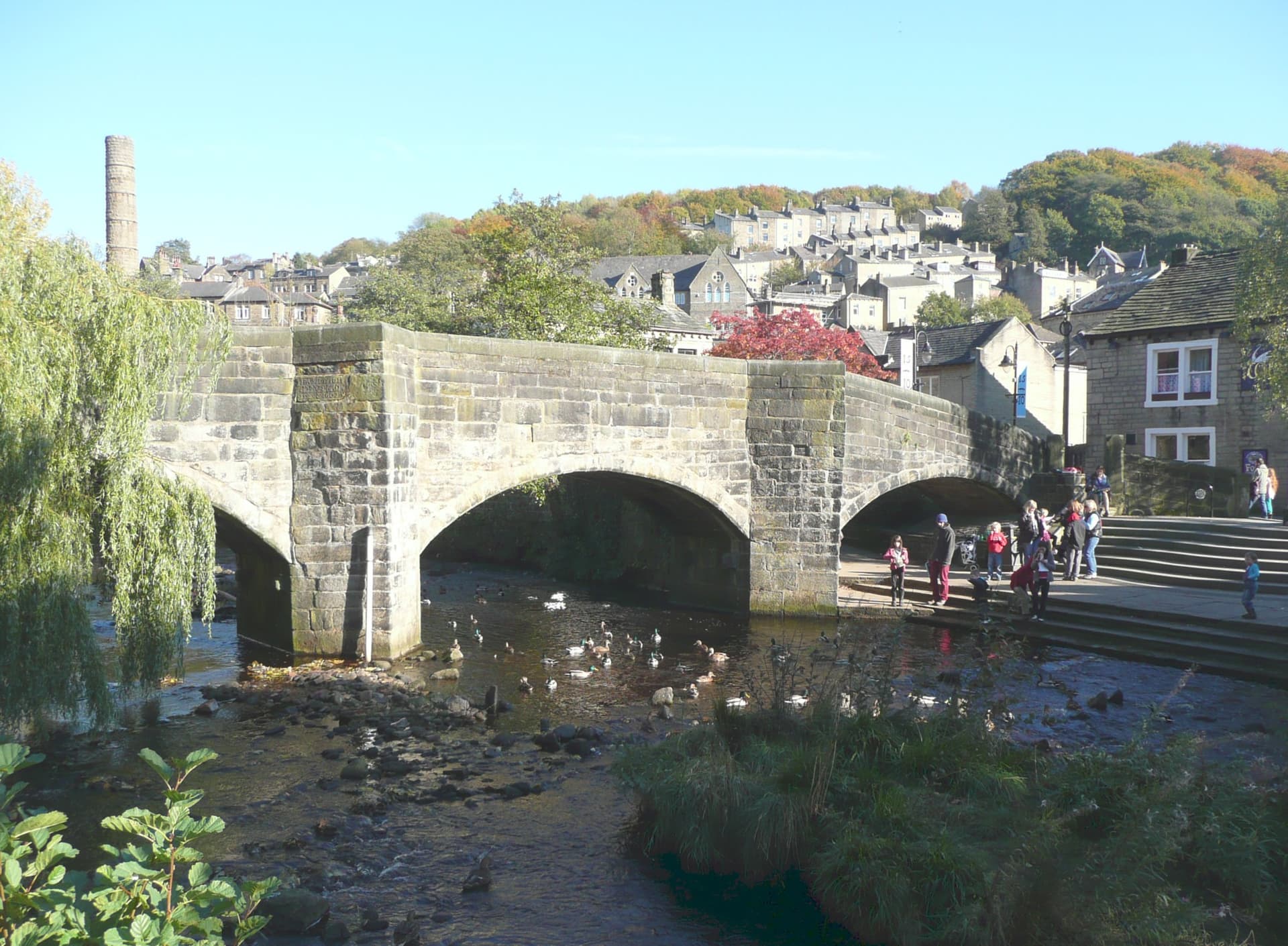 Old stone bridge over a shallow river with ducks, terraced hillside houses and a mill chimney behind