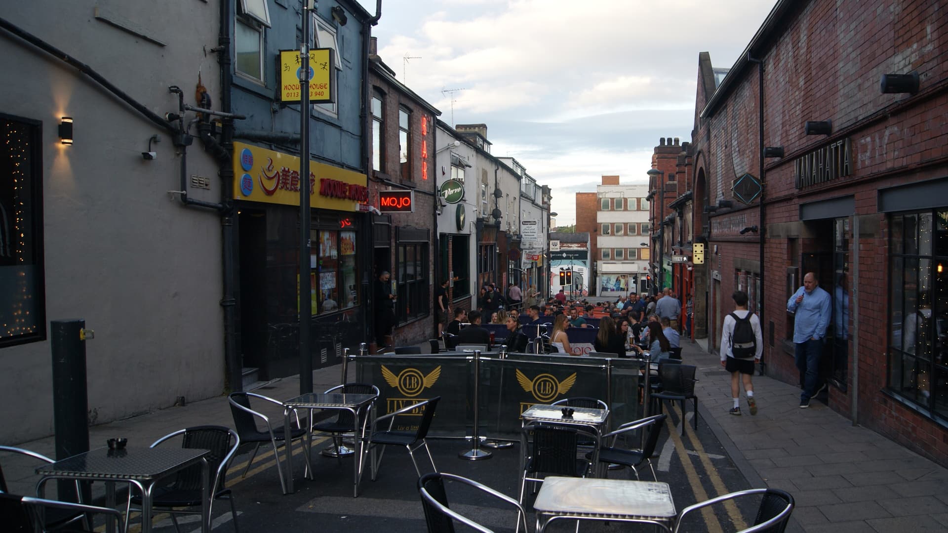 Pavement tables and busy bars along a narrow Leeds street at dusk, with neon signs and brick buildings