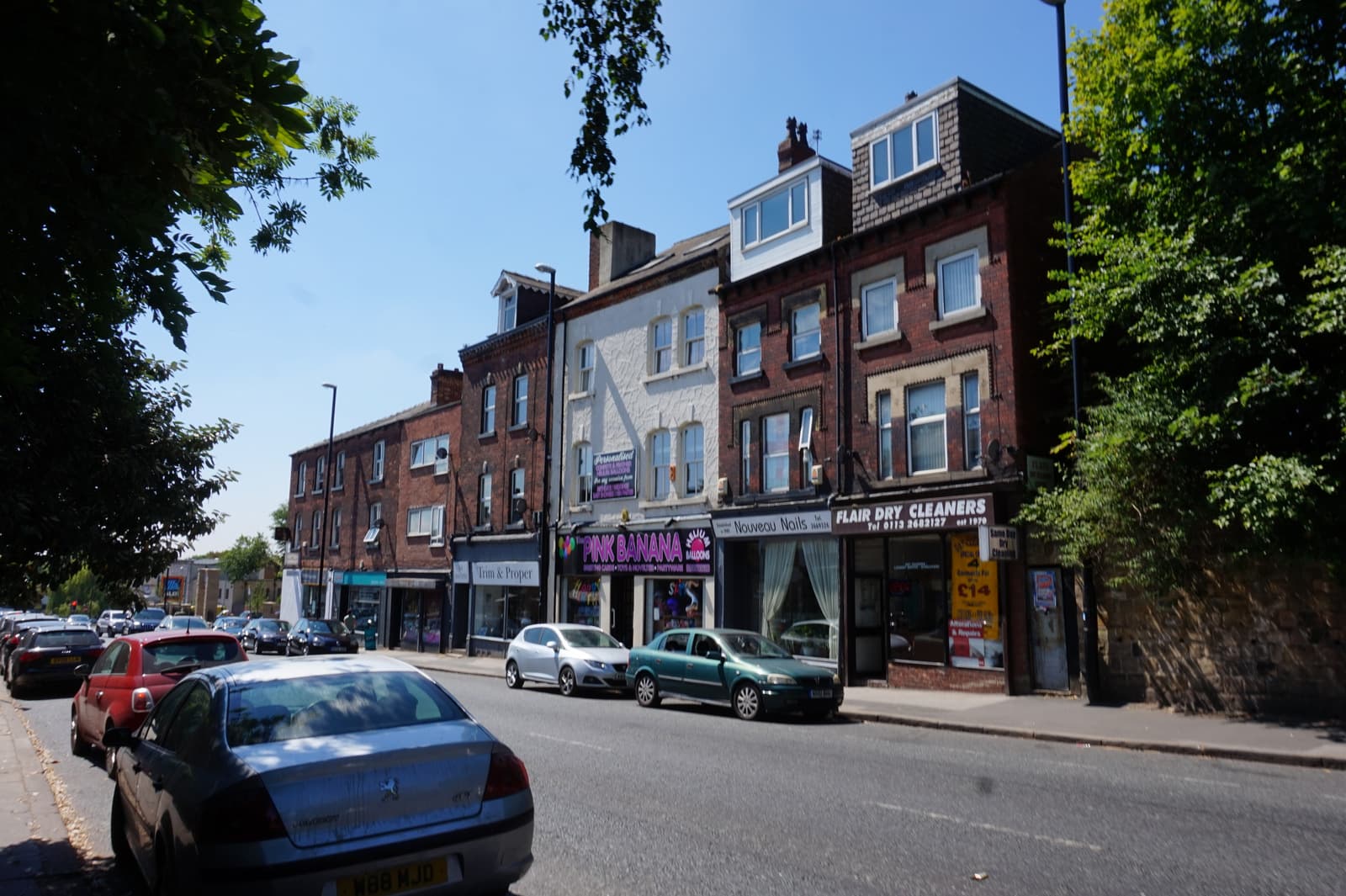 Parked cars along a sunlit street of red-brick terraced shopfronts including a dry cleaner, nail bar and balloon shop